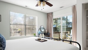 A bedroom with a bed, a ceiling fan, a window with blinds, a balcony with a chair, and a small table with a tray.
