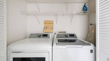 Un cuarto de lavandería con dos lavadoras blancas de carga frontal, un estante de madera y un planchador azul sobre un estante blanco.