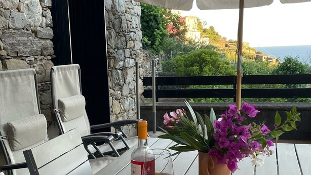 An outdoor seating area with a stone wall, a table with a bottle of red wine, a glass, a basket of lemons, and a potted plant with purple flowers. - Villa Olive Mill Terra Rossa  (Corniglia)
