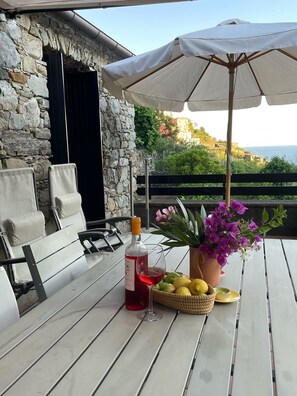 Un espace extérieur aménagé avec des sièges, un mur en pierre, une table sur laquelle se trouve une bouteille de vin rouge, un verre, un panier de citrons et un pot de fleurs avec des fleurs violettes.
