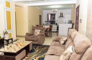 A modern living room with a sofa, armchair, and coffee table. There is a dining area with a table and chairs, and a kitchen in the background. - State House Haven 3 Bedroom Suite (Nairobi)