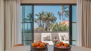 Una mesa de comedor con dos cuencos de fruta en una habitación con ventanas amplias que dan a palmeras y un edificio.