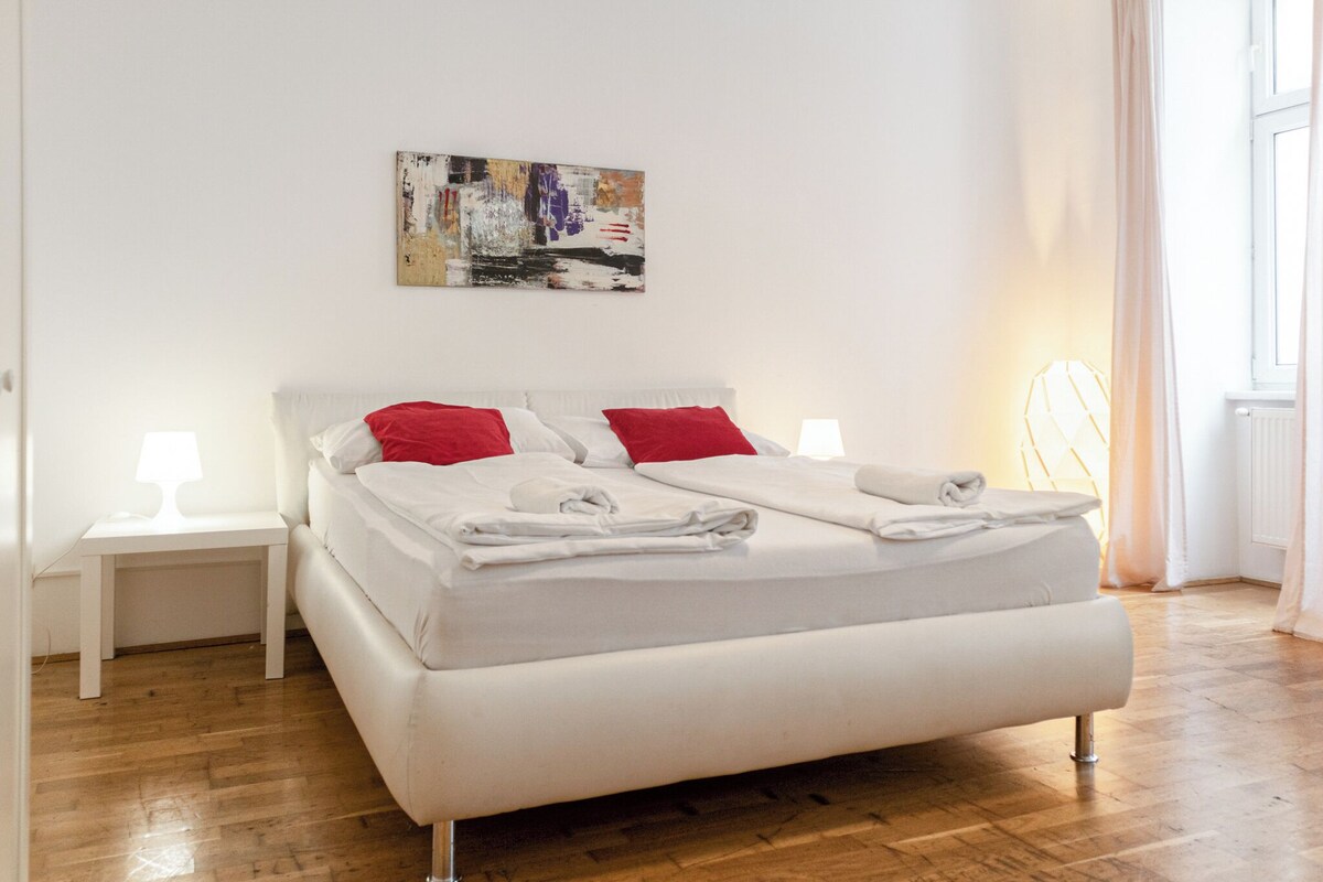 Ein modernes Schlafzimmer mit einem großen Bett, weißer Bettwäsche, roten Kissen und einem kleinen Nachttisch mit Lampe.