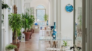 En rymlig korridor med en matplats inredd med vita stolar och ett glasbord, omgiven av stora krukväxter och med utsikt över ett bergigt landskap genom ett stort fönster.