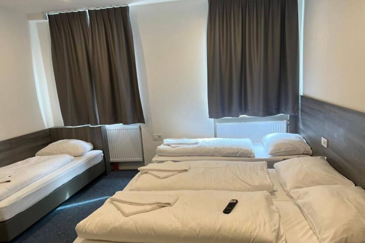 Een hotelkamer met twee eenpersoonsbedden en een tweepersoonsbed, elk met wit beddengoed. Er zijn twee ramen met gordijnen en een radiator is op de muur te zien.