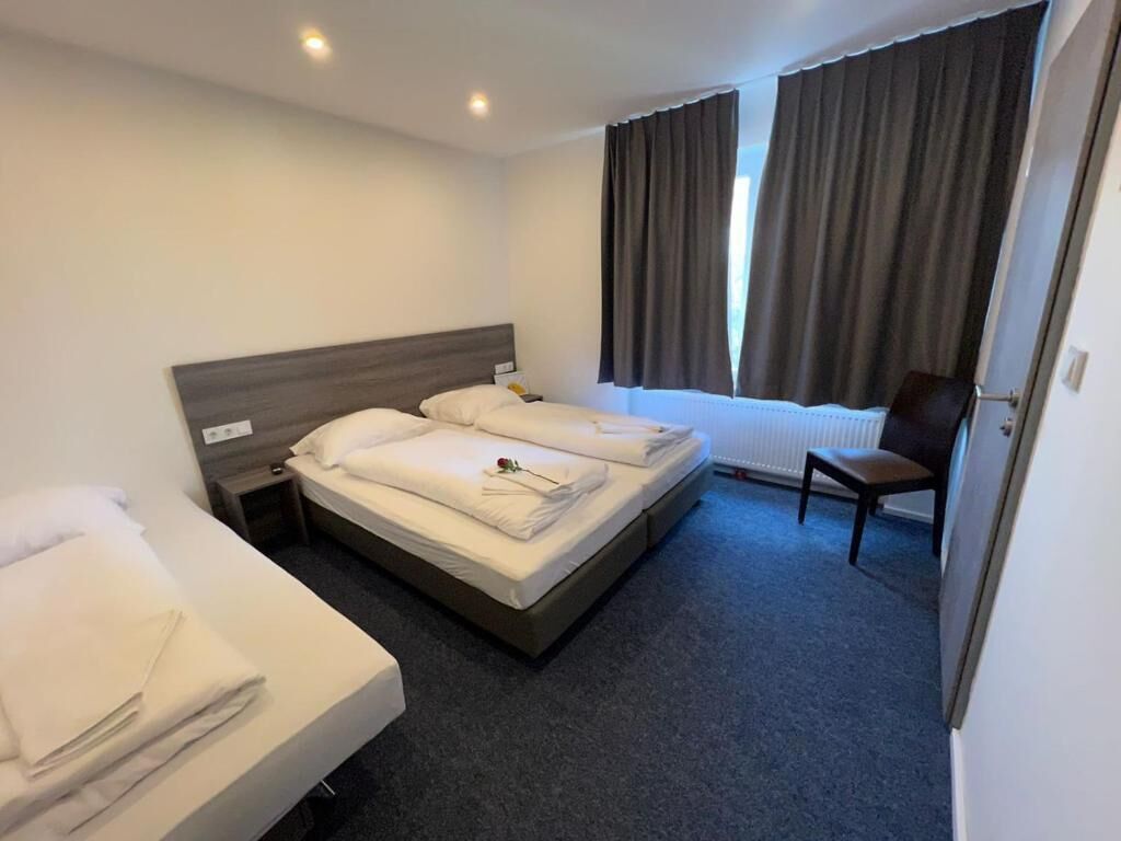 Ein Hotelzimmer mit einem Doppelbett, einem Einzelbett, einem Sessel und einem Fenster mit Vorhängen.