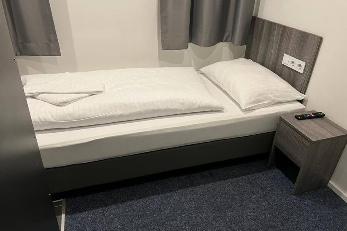Een enkel bed met wit beddengoed, een houten hoofdbord, een klein nachtkastje met een afstandsbediening en een stopcontact aan de muur.