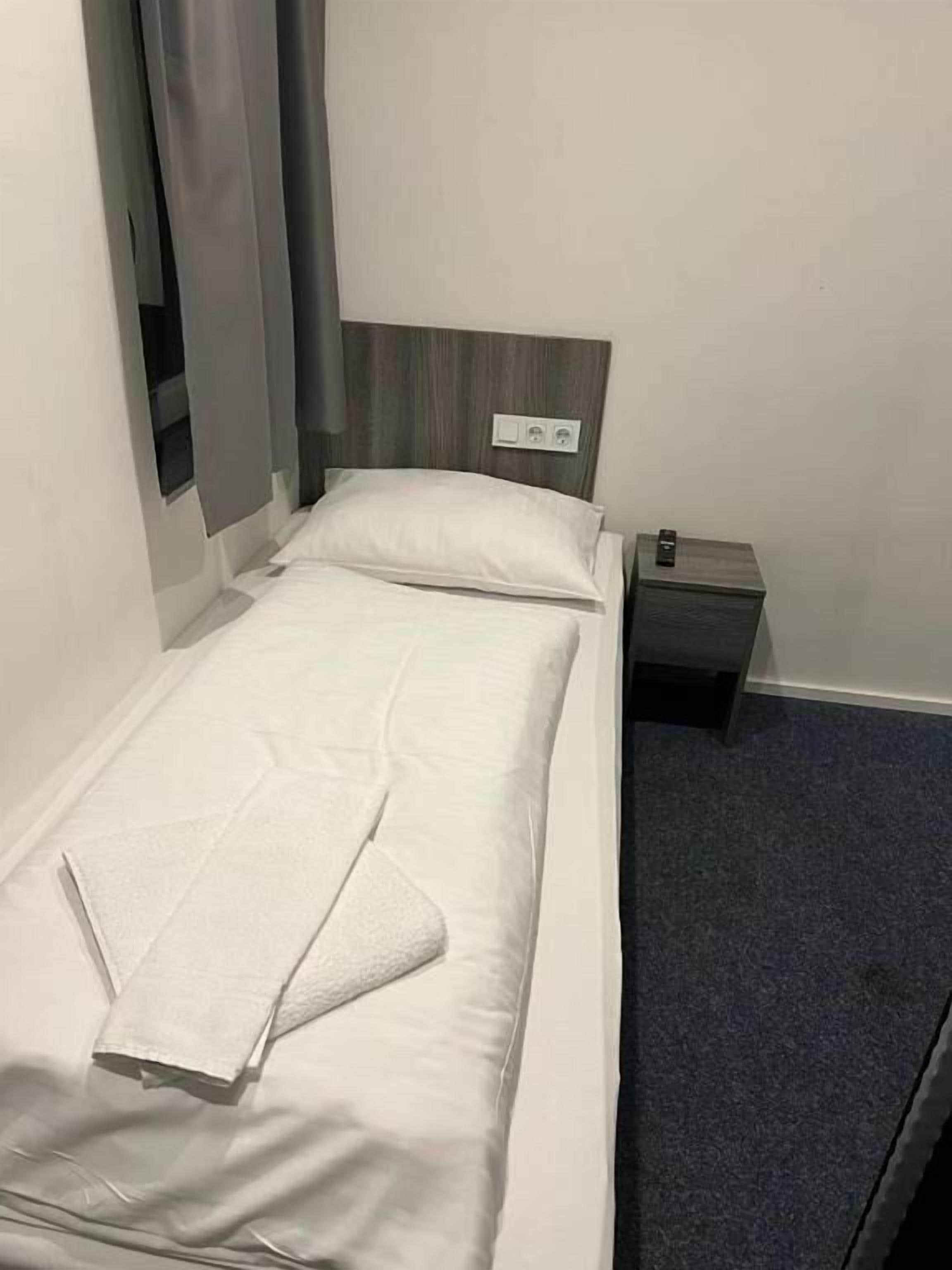 Een eenpersoonsbed met wit beddengoed, een houten hoofdbord, een klein nachtkastje met een lamp en een donker vloerkleed.