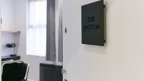 Een kamer met een raam, een radiator, een stoel, een bureau met een lamp, en een bordje met de tekst 'THE HATTON'.