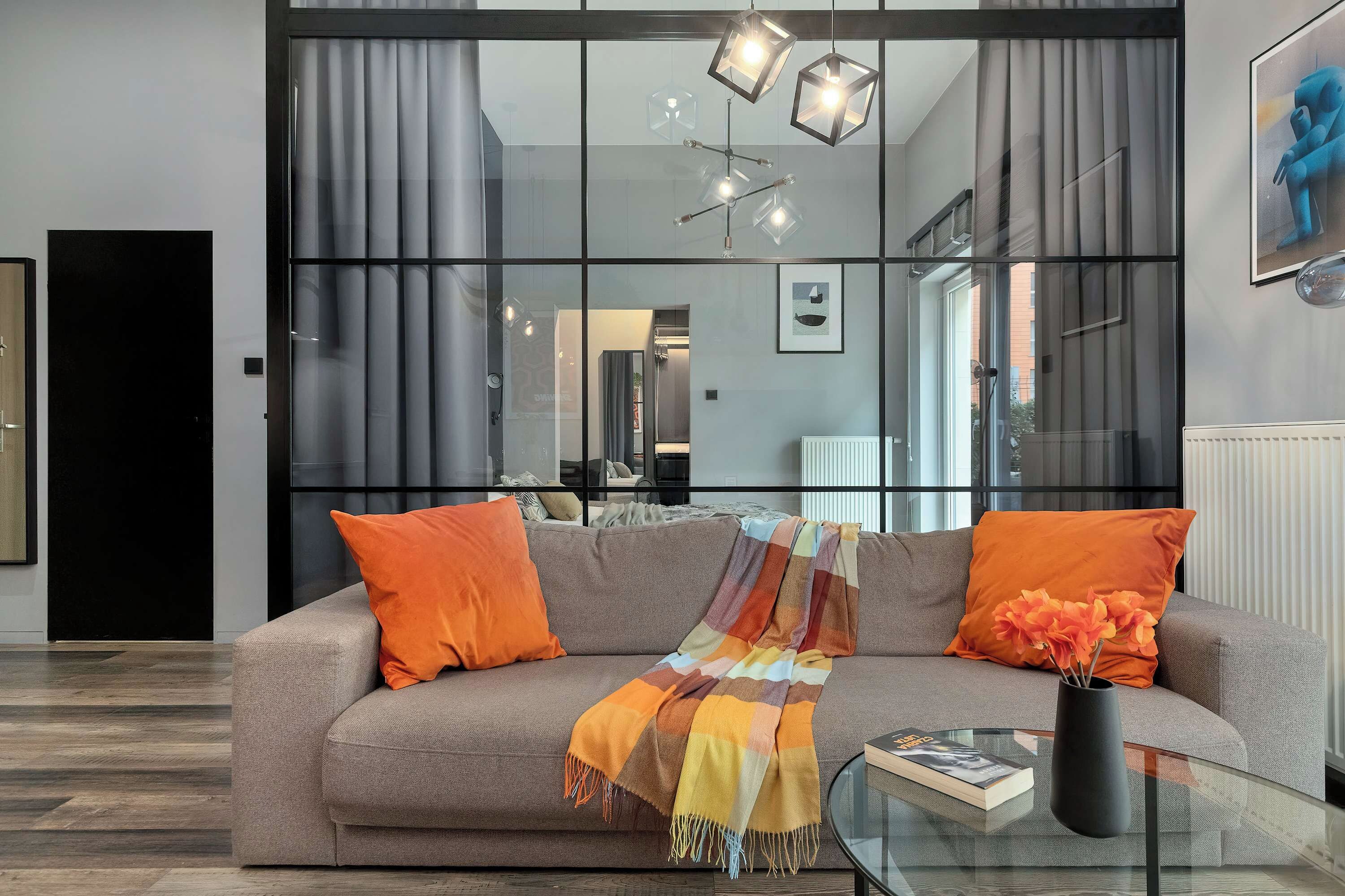 A modern living room with a sofa, a glass coffee table, and a book. There is a window with curtains, a painting on the wall, and a radiator.