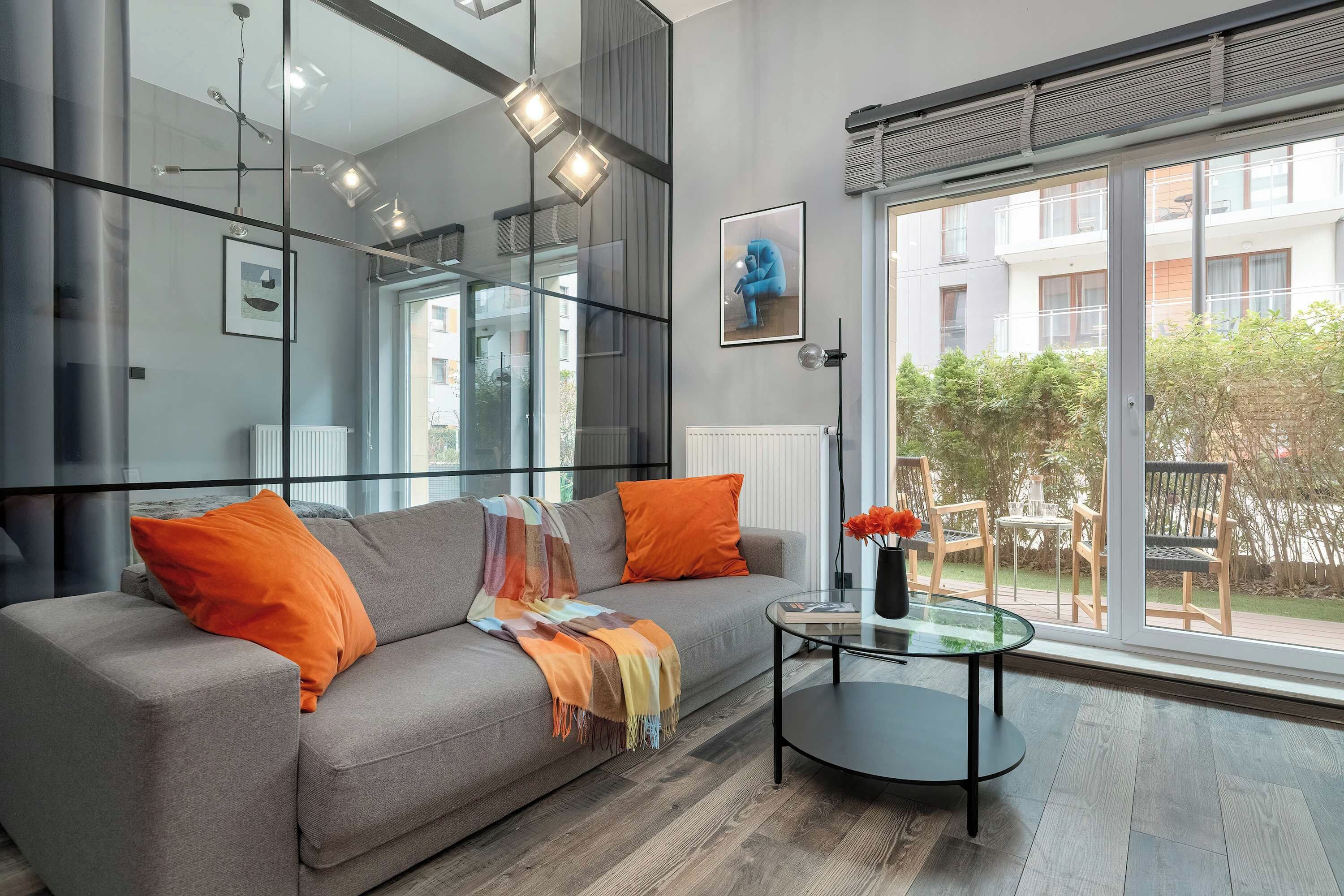 Ein modernes Wohnzimmer mit einem grauen Sofa, orangefarbenen Kissen, einem Glastisch und einem großen Fenster mit Blick auf eine Wohngegend.