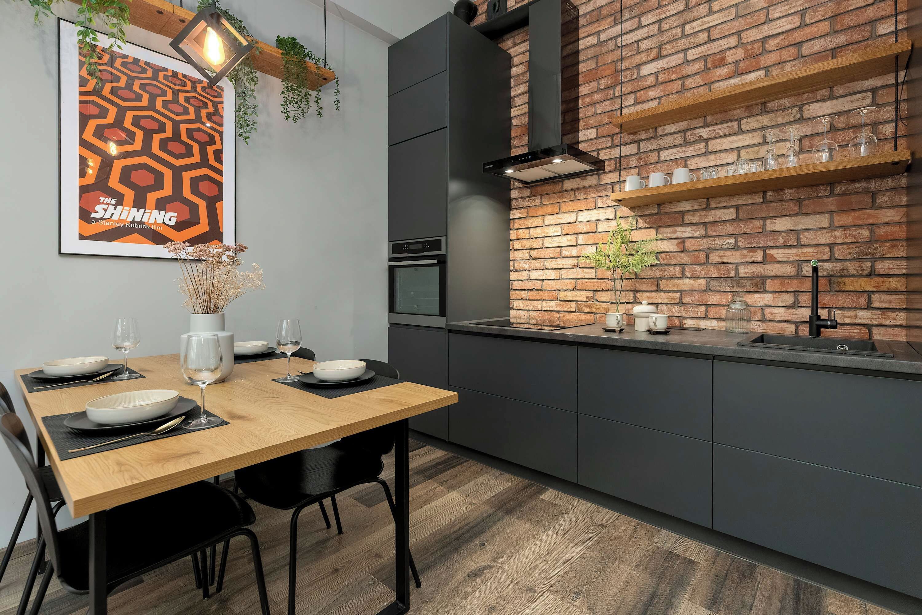 A modern kitchen with a brick wall, a dining table set with plates and glasses, and a hanging light fixture.