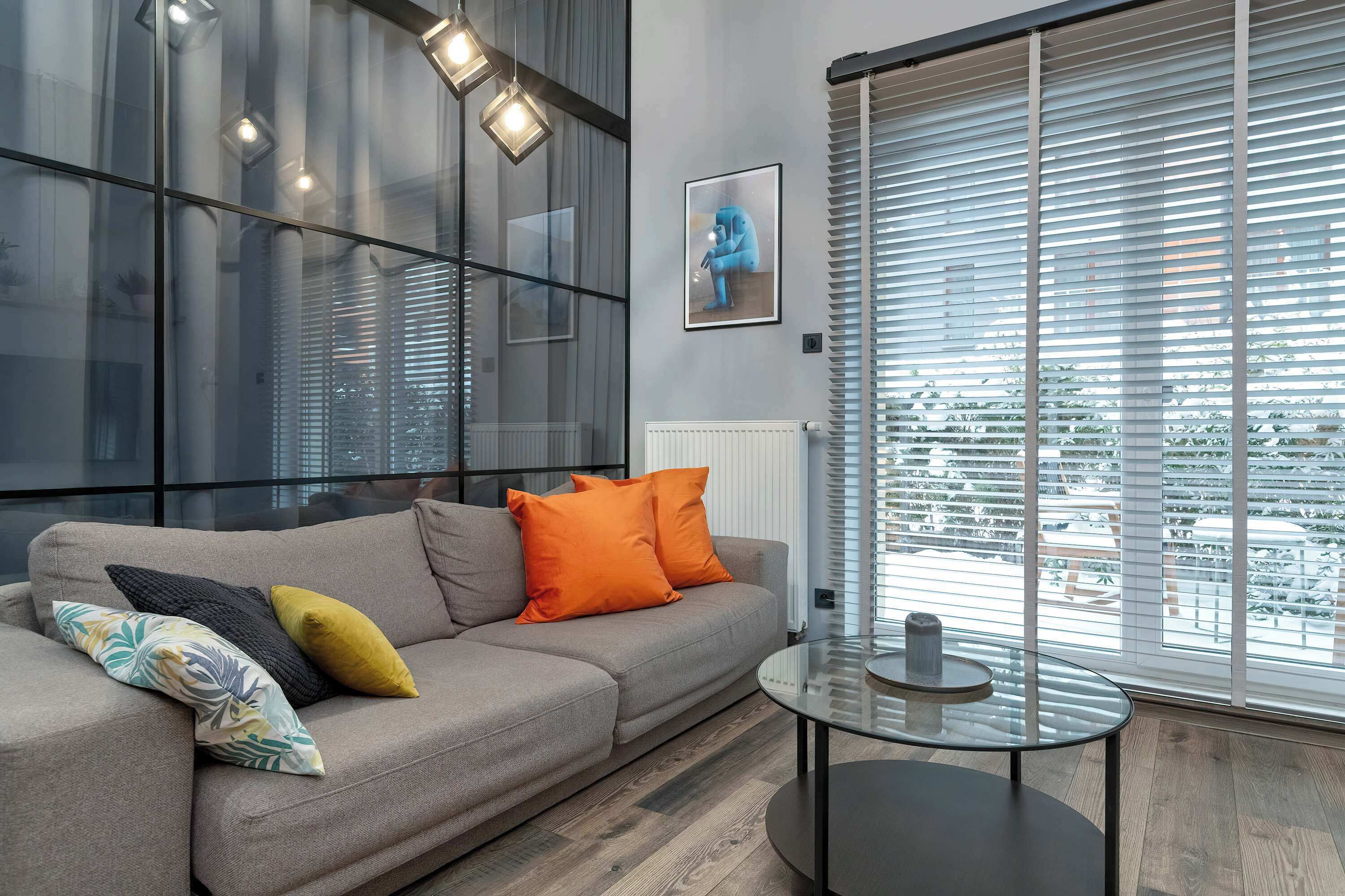 Un salón moderno con un sofá gris, cojines naranja y estampados, una mesa de centro de vidrio y vistas a la ciudad a través de grandes ventanales.