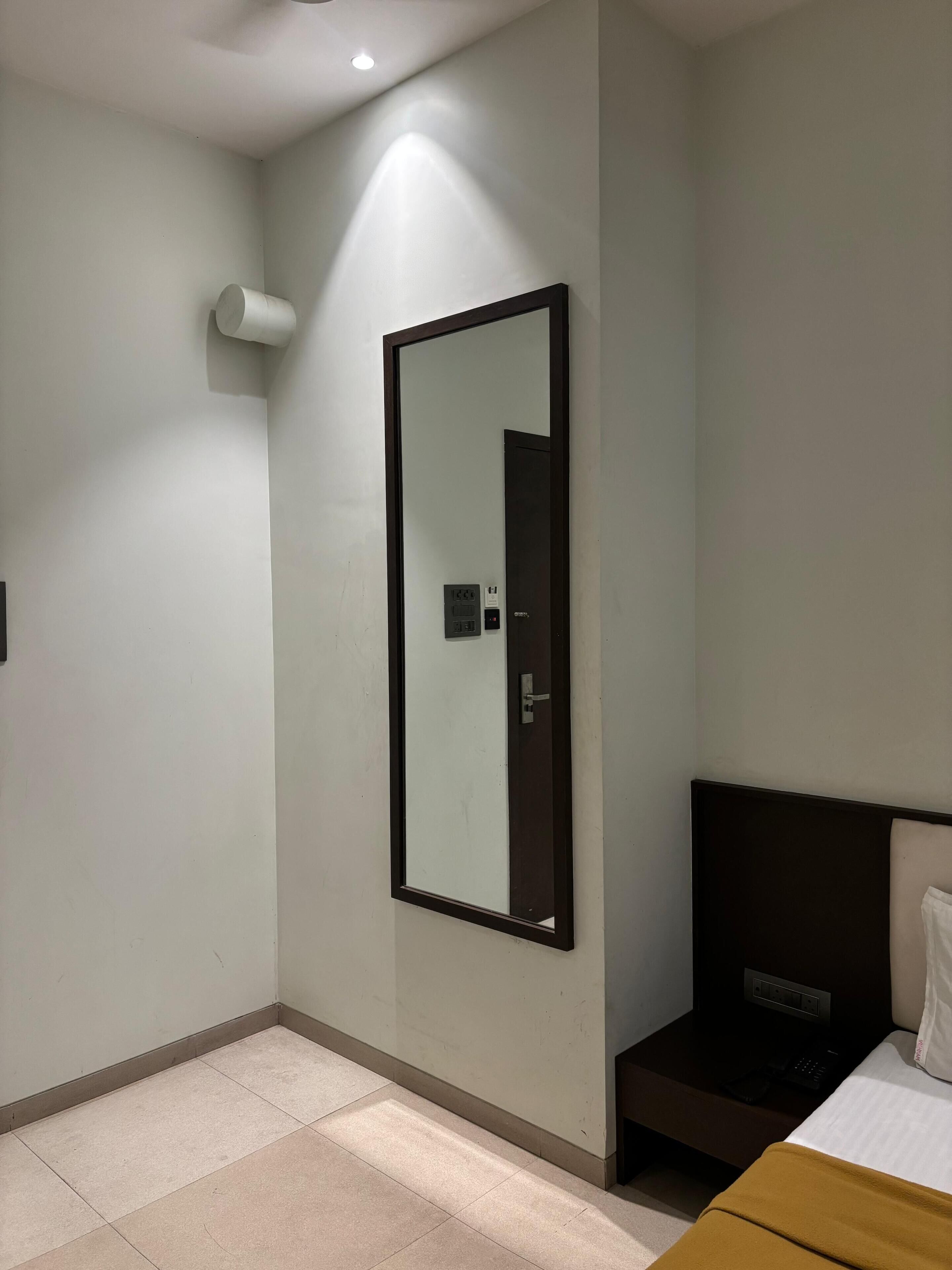 A hotel room with a bed, a nightstand, a mirror, and a wall-mounted control panel.