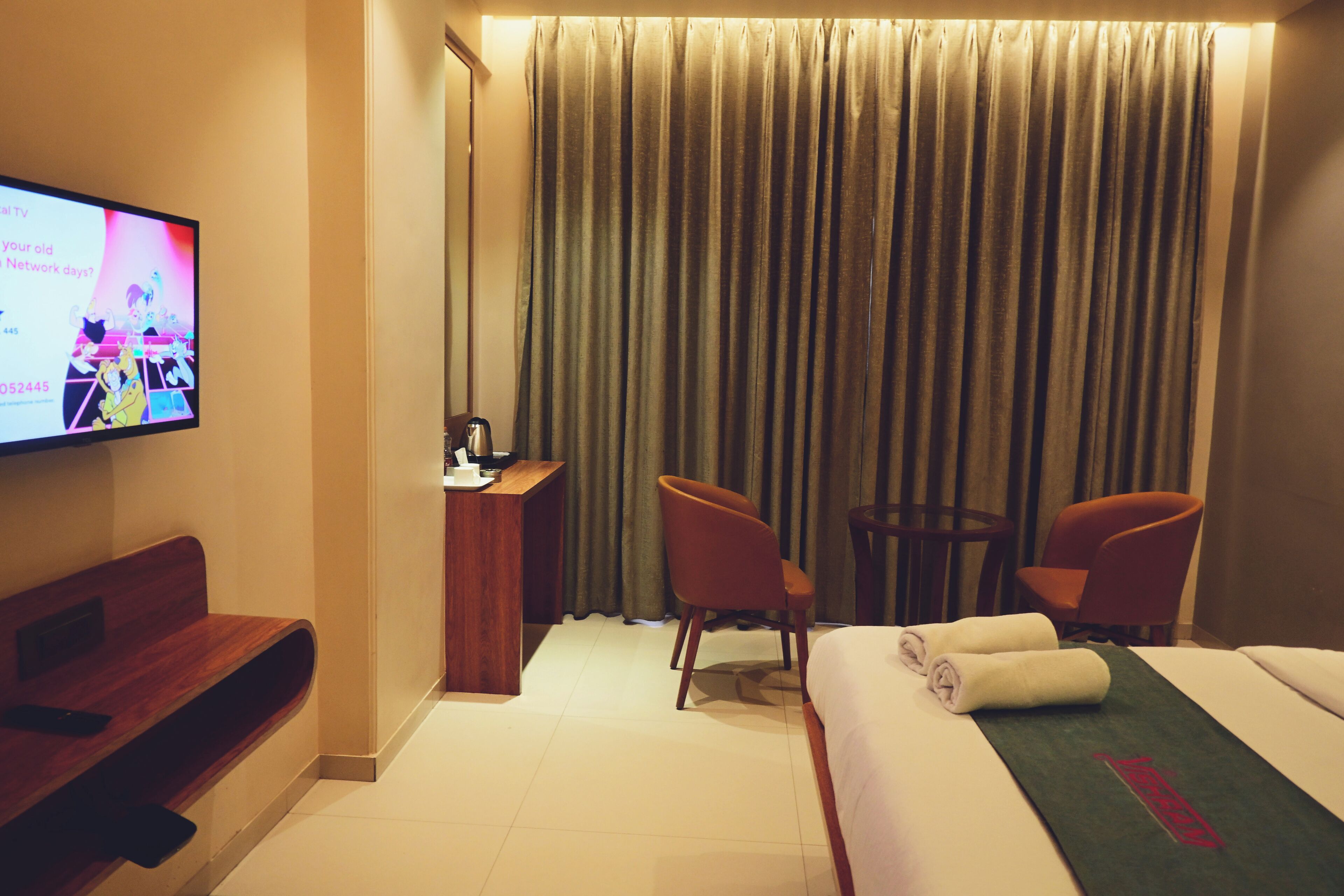 A hotel room with a bed, two chairs, a desk, a TV, and a curtain.