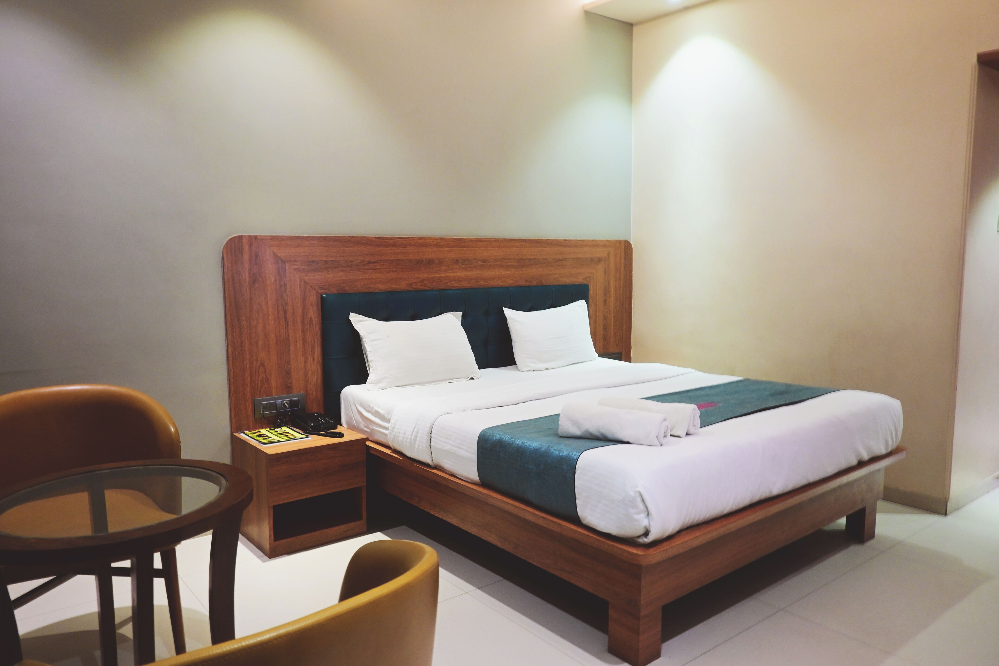 A neatly made bed with white linens and a wooden headboard, a small wooden nightstand with a phone and a book, and two chairs in a corner.