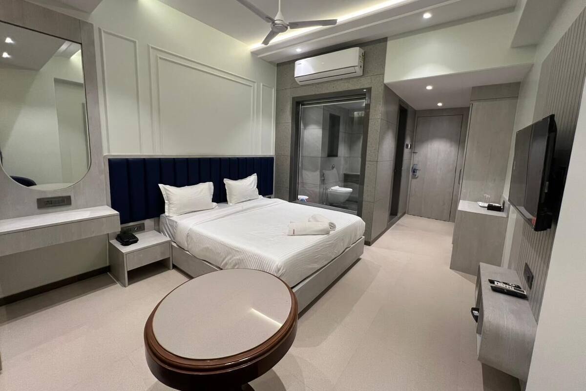 A modern hotel room with a bed, bedside tables, a round table, a ceiling fan, an air conditioning unit, a TV, and a bathroom with a glass shower enclosure.