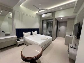 A modern hotel room with a bed, bedside tables, a round table, a ceiling fan, an air conditioning unit, a TV, and a bathroom with a glass shower enclosure. - Vishram Executive Rooms (Vasai)