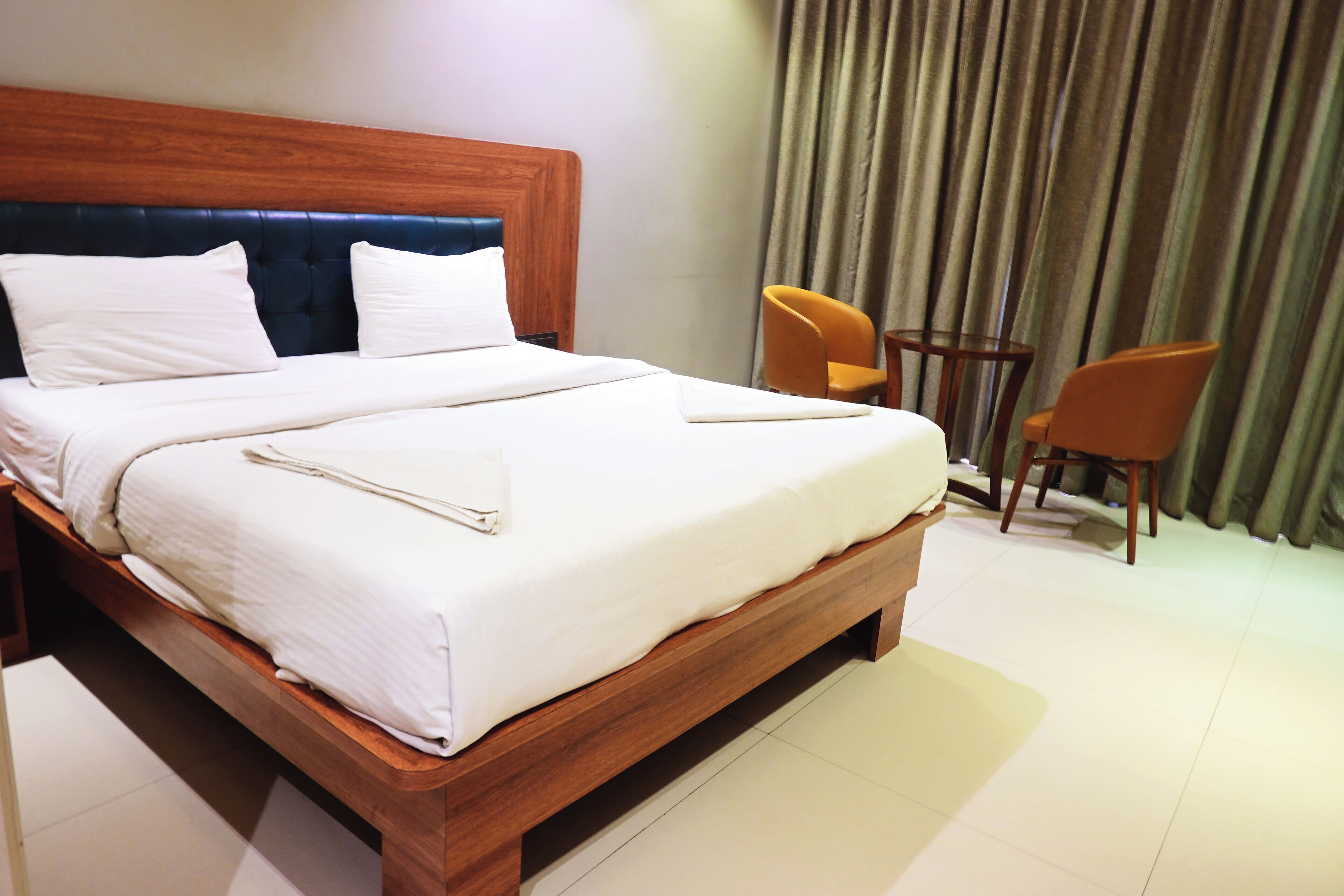 A neatly made bed with white linens and a wooden headboard, two orange chairs, and a large window with curtains.