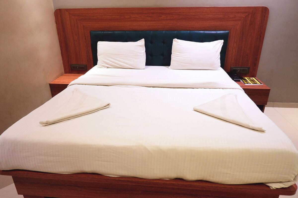 A neatly made bed with white linens and two pillows, a wooden headboard, and a bedside table with a remote control and a book.
