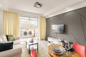 A modern living room with a sofa, dining table, and chairs, a flat-screen TV mounted on the wall, and a balcony with yellow curtains. - Cozy 1-bedroom apartment with AC in serene Nairobi (Nairobi Kileleshwa)