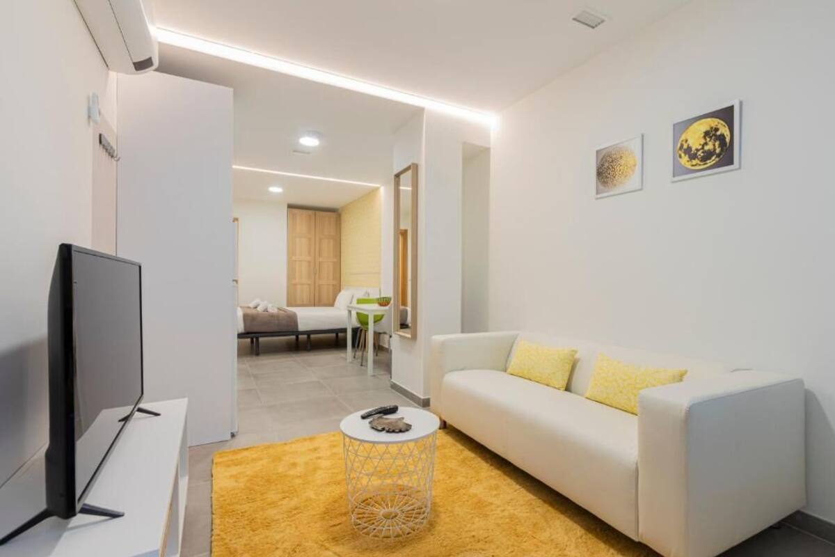 Een moderne woonkamer met een flatscreen televisie, een witte bank met gele kussens, een ronde salontafel en kunst aan de muur.