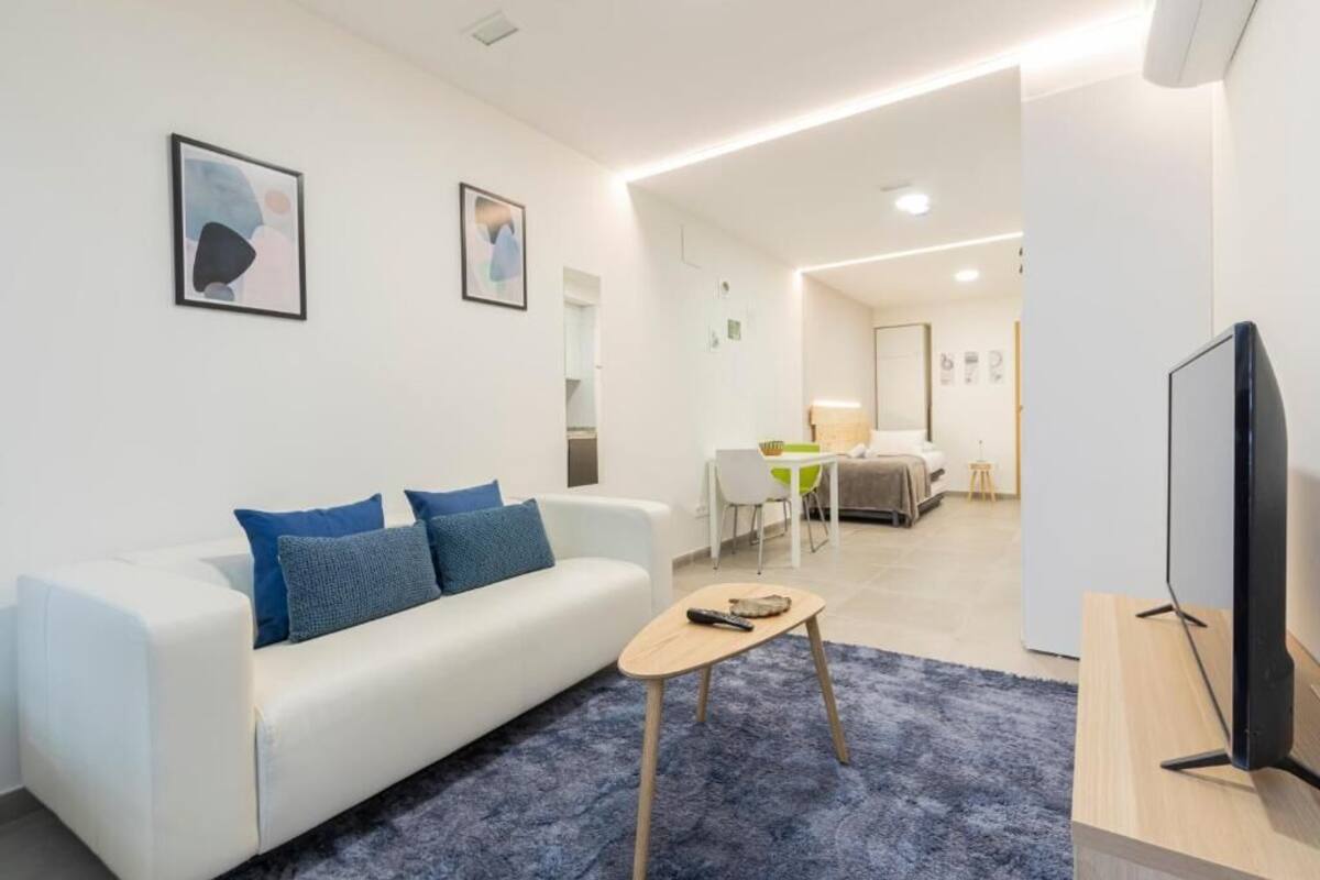Een moderne woonkamer met een witte bank, blauwe kussens, een klein houten salontafel en een flatscreen televisie op een houten standaard.