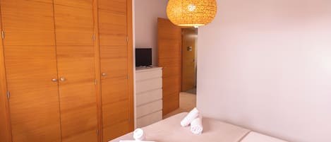 A bedroom with wooden wardrobes, a bed with towels, a hanging light fixture, and a television in the background.