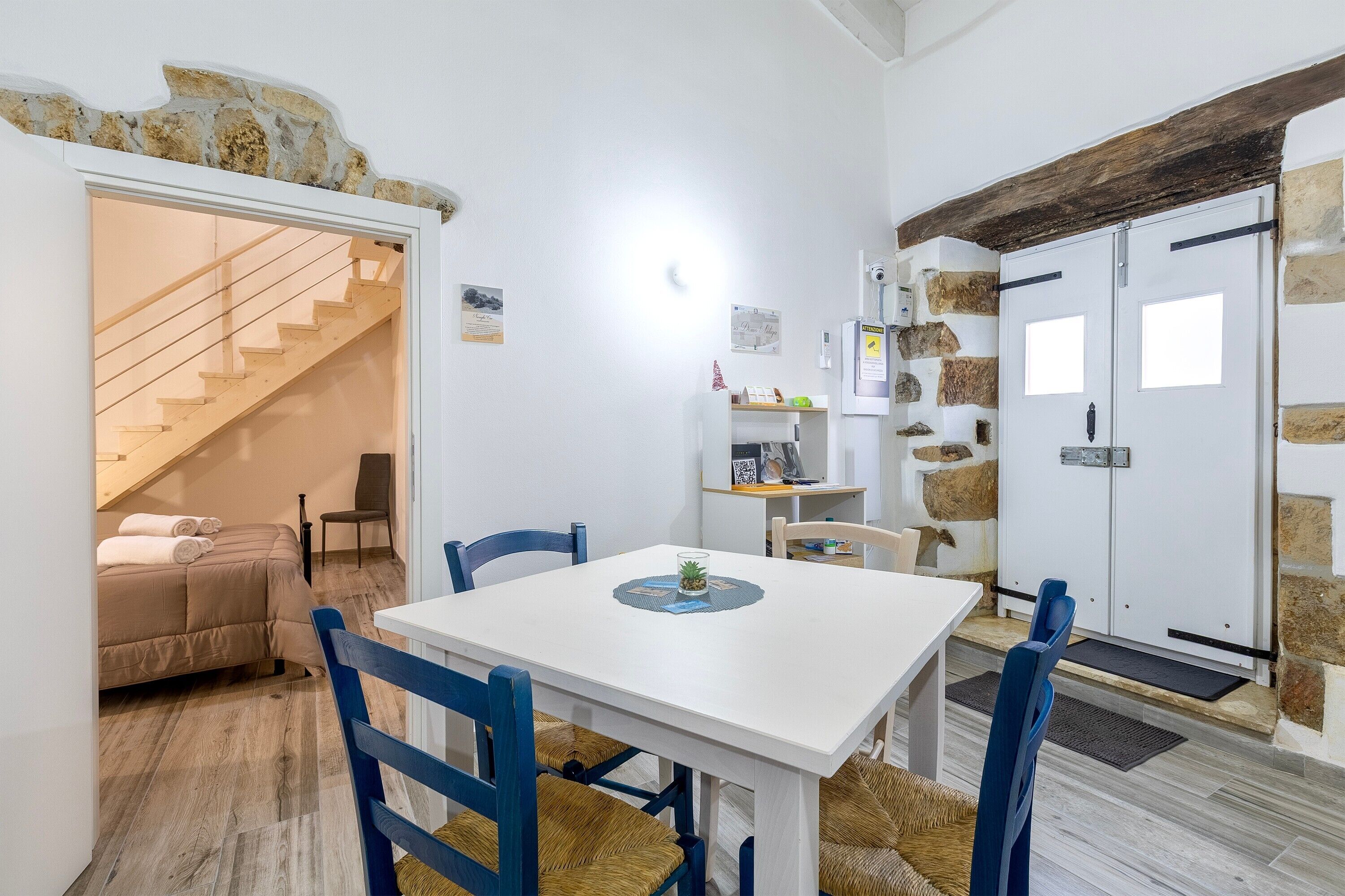 A room with a white table, blue chairs, a bed with a wooden headboard, a staircase, and stone walls.