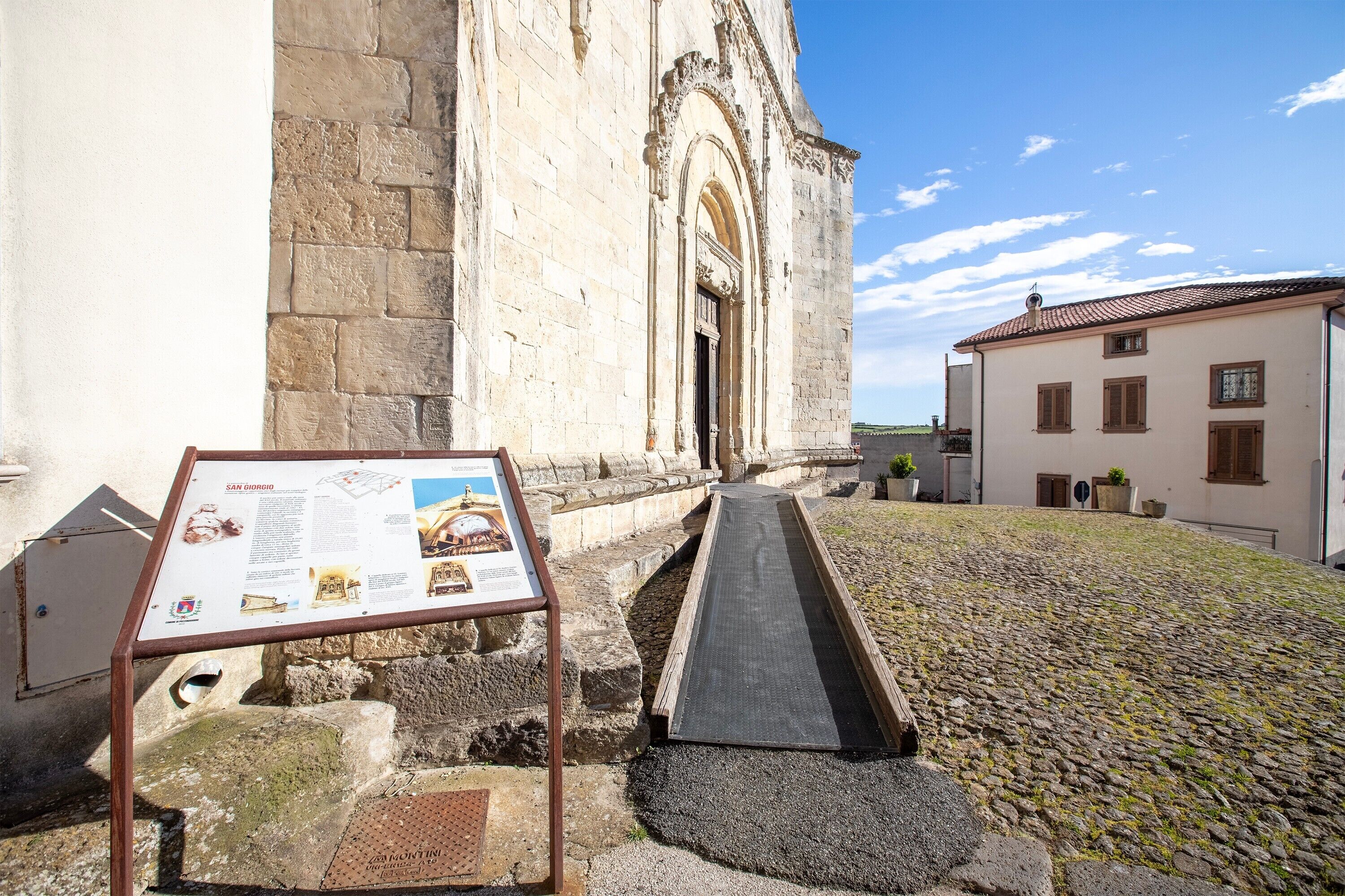 Un bâtiment en pierre avec un chemin pavé menant à l’entrée, un panneau d’information historique et un sol en pavés.
