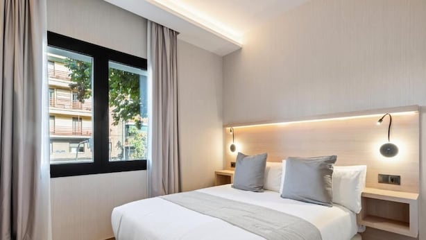 A modern hotel room with a large bed, a nightstand, a built-in shelf, and a window with curtains. - Dobo Madrid Ricardos (Madrid)