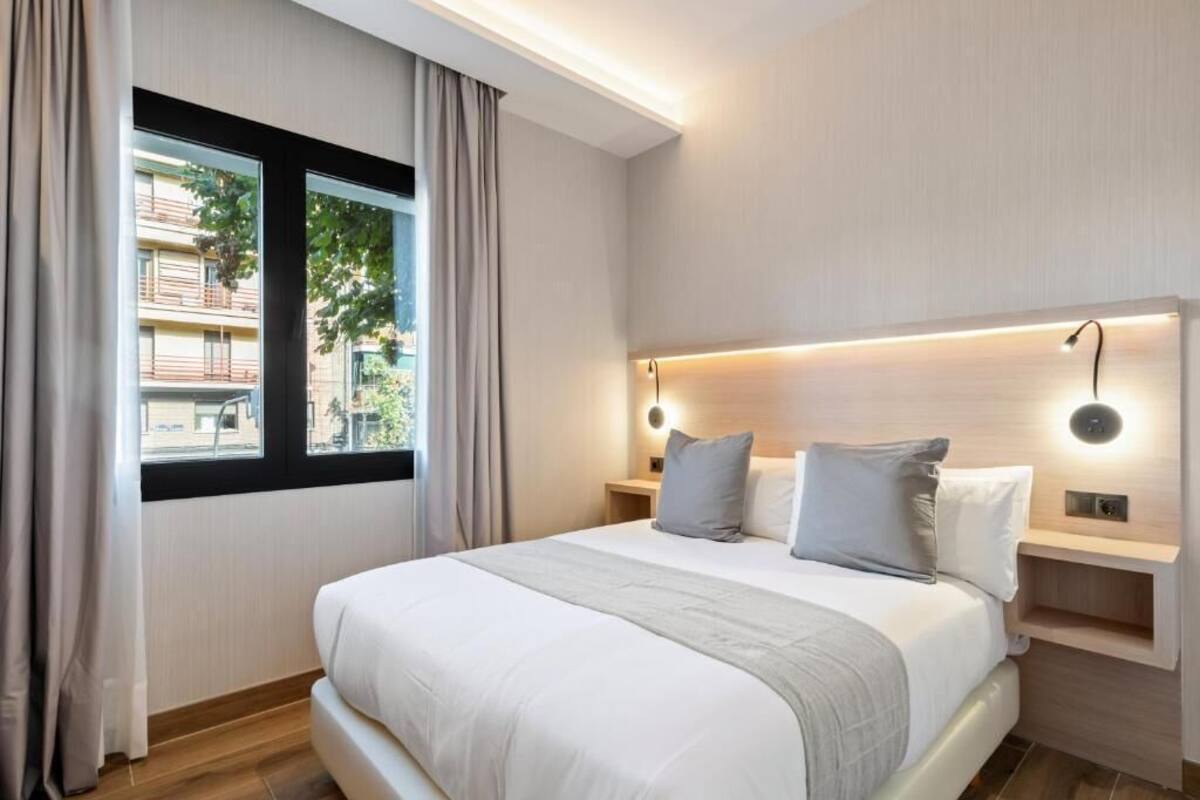 A modern hotel room with a large bed, a nightstand, a built-in shelf, and a window with curtains.