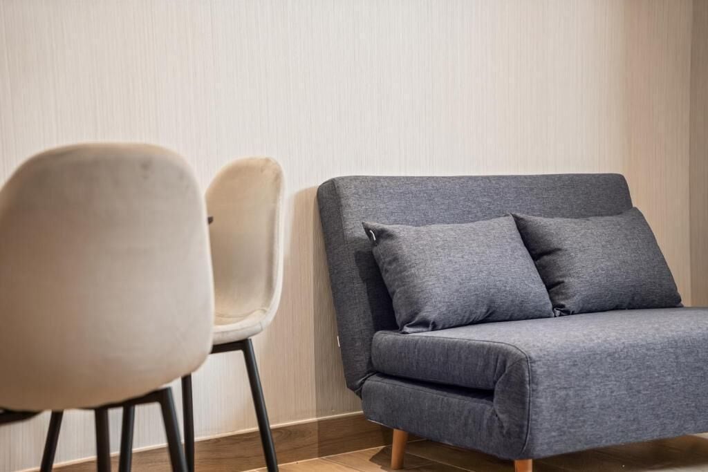 A grey sofa with two cushions and a white chair in a room with a wooden floor and light-colored walls.