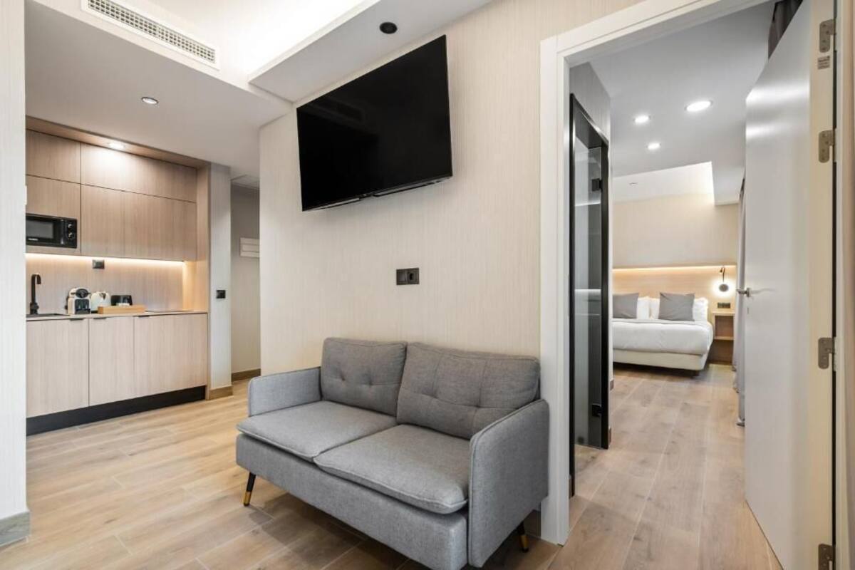 A modern living room with a grey sofa, a flat-screen TV mounted on the wall, and a kitchen area with wooden cabinets and a microwave.