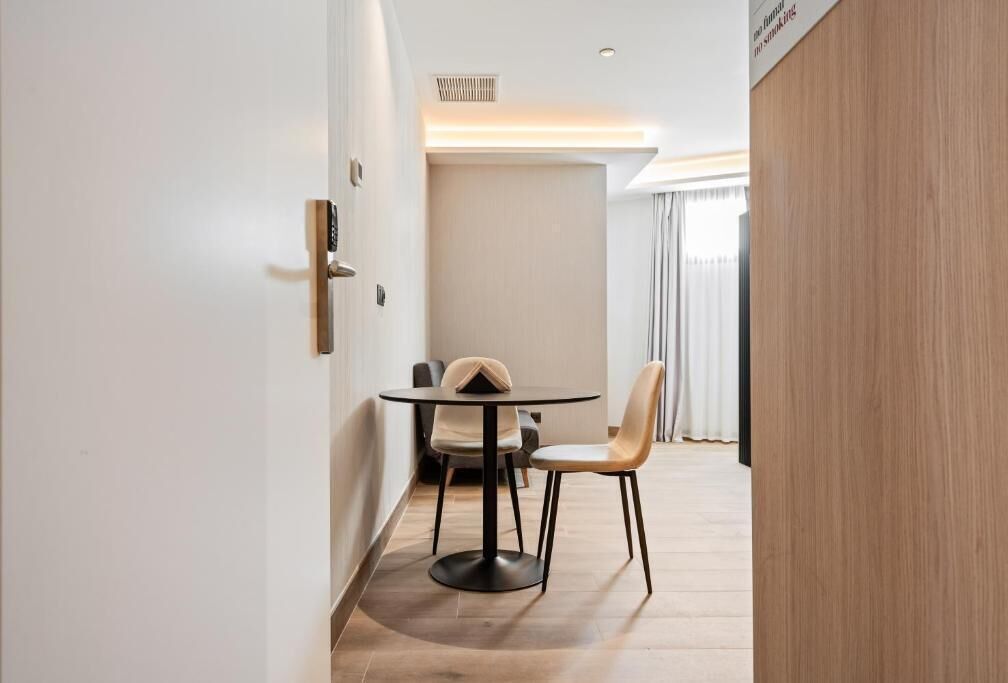 A modern hotel room with a round table and two chairs, a wooden floor, and a view of the outdoors through a window with curtains.