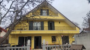 A two-story yellow house with a balcony, a statue, and a small garden area. - Beautiful Chalet-style house. 10 min from Paris (Colombes)