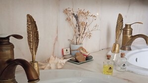 A bathroom with a white sink, two brass-feathered faucets, a potted plant, and two bottles of shampoo. - Beautiful Chalet-style house. 10 min from Paris (Colombes)