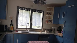 A compact kitchen with blue cabinets, a red checkered table, a window with lace curtains, and a hanging light fixture. - Beautiful Chalet-style house. 10 min from Paris (Colombes)