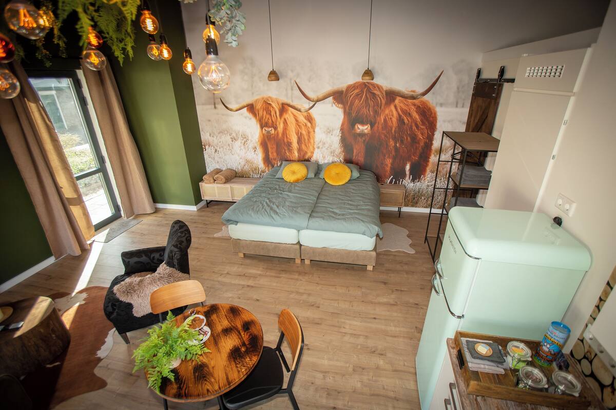 Un dormitorio con cama, una mesa de comedor con sillas, una pequeña cocina y un mural grande de ganado de alta montaña.