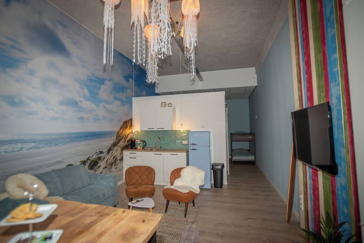 Sala de estar con un mural de playa, una mesa de comedor de madera, dos sillones, una pequeña cocina y un televisor.