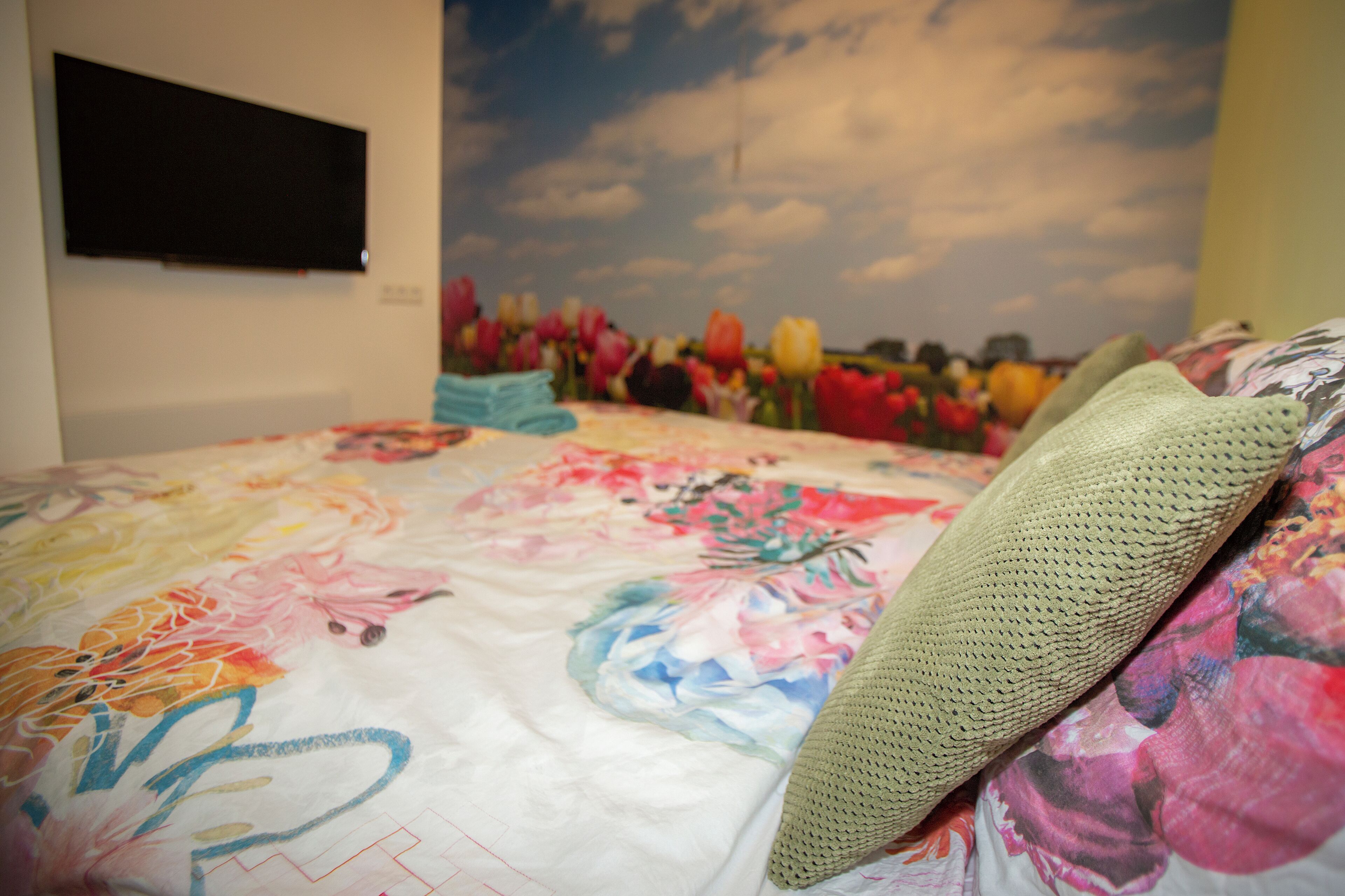 Una cama con una colcha estampada con motivos florales, un televisor instalado en la pared y un mural grande con diseño floral de fondo.
