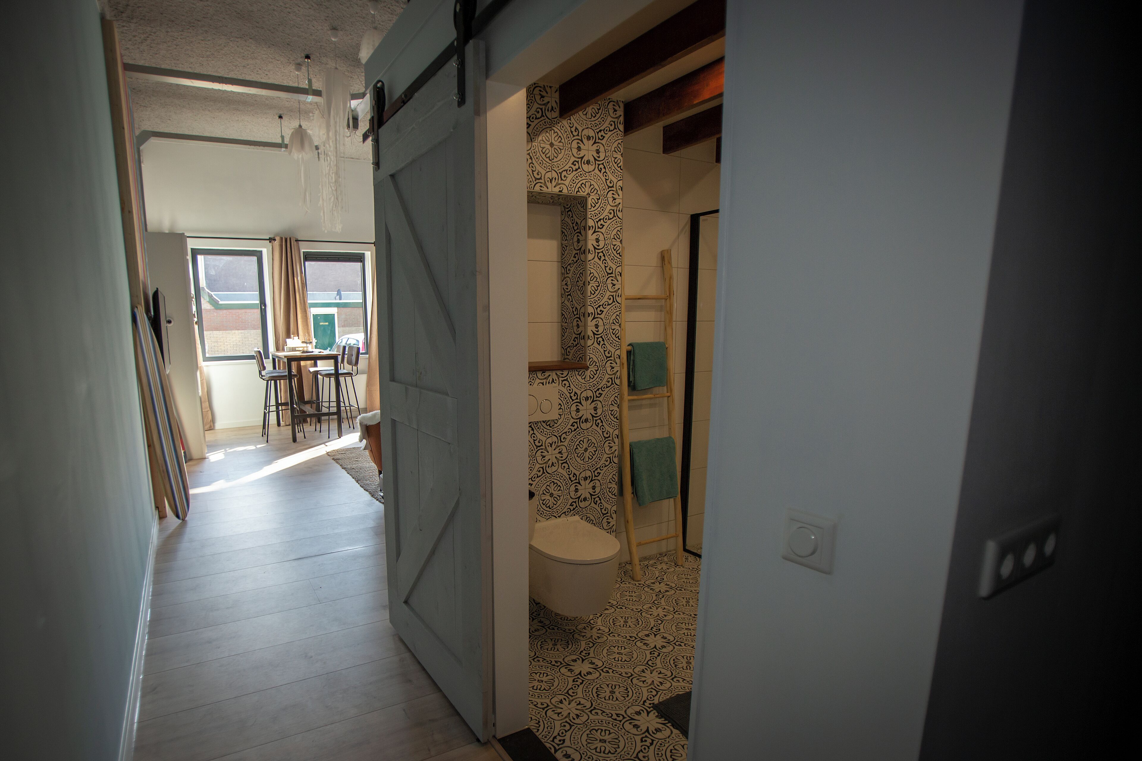A hallway leading to a bathroom with patterned tiles, a toilet, and a towel rack.
