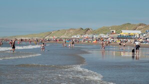 Une plage bondée où les gens pratiquent diverses activités, une rive sablonneuse et un bâtiment avec un drapeau en arrière-plan.