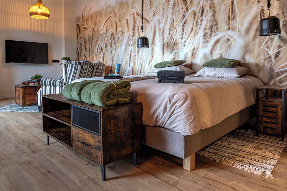 Un dormitorio moderno con una cama grande, una consola de madera, un televisor montado en la pared y una pared decorativa con hierbas altas.