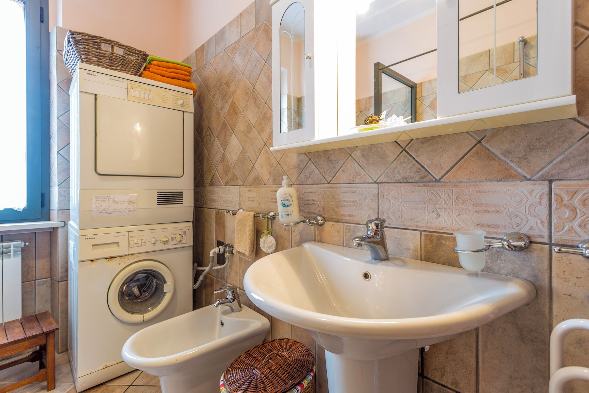 A compact bathroom with a washing machine, bidet, sink, and a small wicker stool.