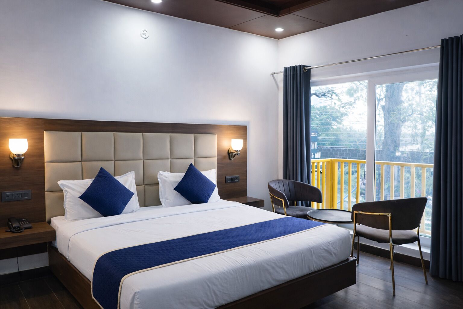 Habitación de hotel con una cama grande, dos mesitas de noche, una silla, una mesita pequeña y un balcón con cortinas.
