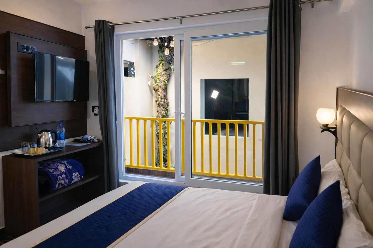 A hotel room with a bed, a TV, a desk with a kettle, and a balcony with yellow railings.