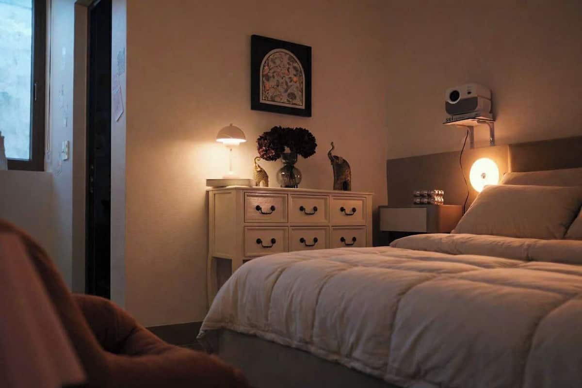 Un dormitorio con cama, lámparas de noche, mesita de noche, un jarrón decorativo y un proyector montado en la pared.