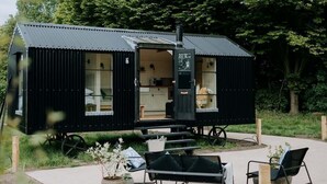 A compact, black metal cabin with a kitchen area visible through an open door, surrounded by outdoor seating and potted plants. - Elsham Hall Bothies: Countryside Retreat with Garden Views & Free Park Access (Elsham)