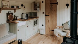 A cozy kitchen with wooden cabinets, a built-in oven, and a coffee machine. There's a wooden bench with cushions, a small table with a kettle, and a black heater. - Elsham Hall Bothies: Countryside Retreat with Garden Views & Free Park Access (Elsham)